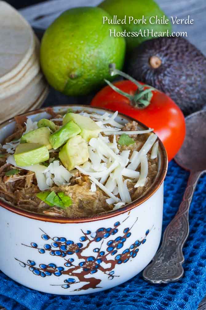 A bowl Pulled Pork Green Chile Verde surrounded by fresh lime, tomato, avocado, and tortillas.