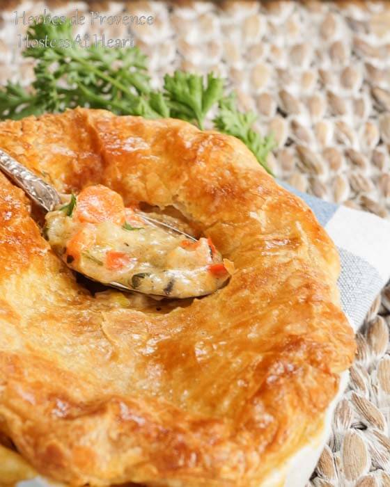 A pot pie topped with a puff pastry crust sitting in a white dish over a blue-checked napkin.  A spoon of the filling hovers over the pie.