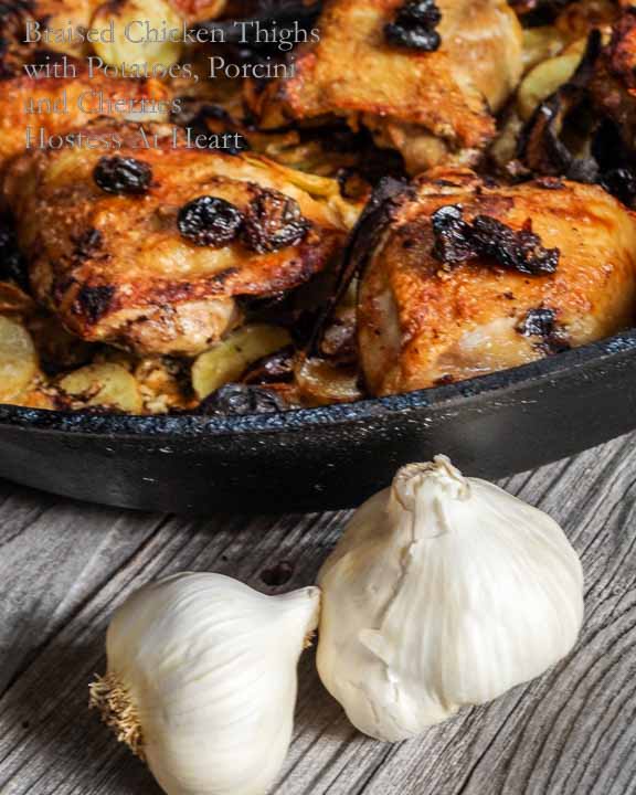 Cast Iron pan filled with cooked chicken thighs with potatoes, Porcini & dried cherries. Two bulbs of garlic sit in the front.