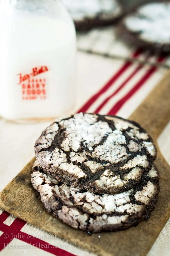 A stack of Dark Chocolate Cookies sitting in front of a bottle of milk and a cooling rack full of cookies.A close up of a piece of cake sitting on top of a table