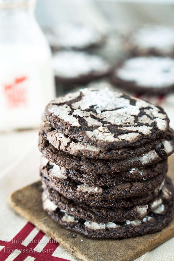A stack of Dark Chocolate Cookies sitting in front of a bottle of milk and a cooling rack full of cookies.