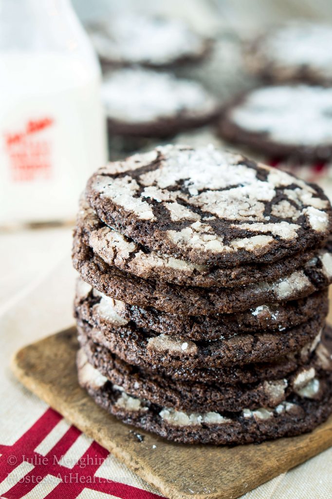 A stack of Dark Chocolate Cookies sitting in front of a bottle of milk and a cooling rack full of cookies.