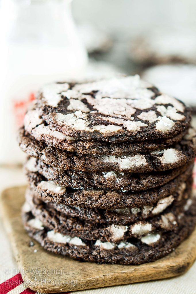 A stack of Dark Chocolate Cookies sitting in front of a bottle of milk and a cooling rack full of cookies.A piece of chocolate cake on a plate, with Cookie and Chewy