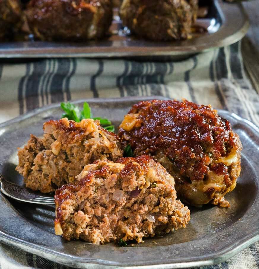 A metal plate filled with a large meatball and one meatball cut in half garnished with fresh parsley over a striped napkin. A pan of meatballs sits in the background.