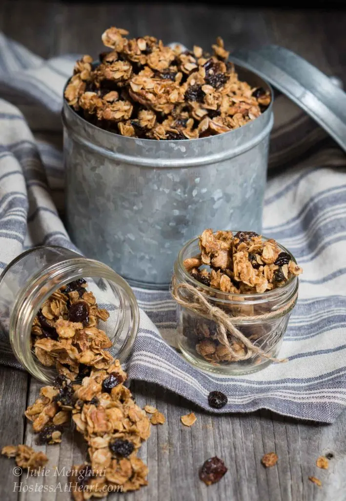 A metal tin filled with Vanilla Nut Granola sitting on a blue striped towel. Jars filled with the granola sit in front of the tin with one of the jars tipped on its side with granola spilling out.