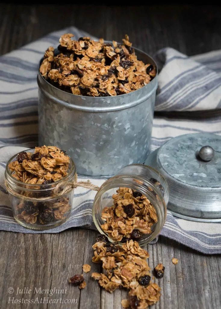 A large tin of granola with a small jar filled in the front and a jar sitting on its side with granola spilling from it.