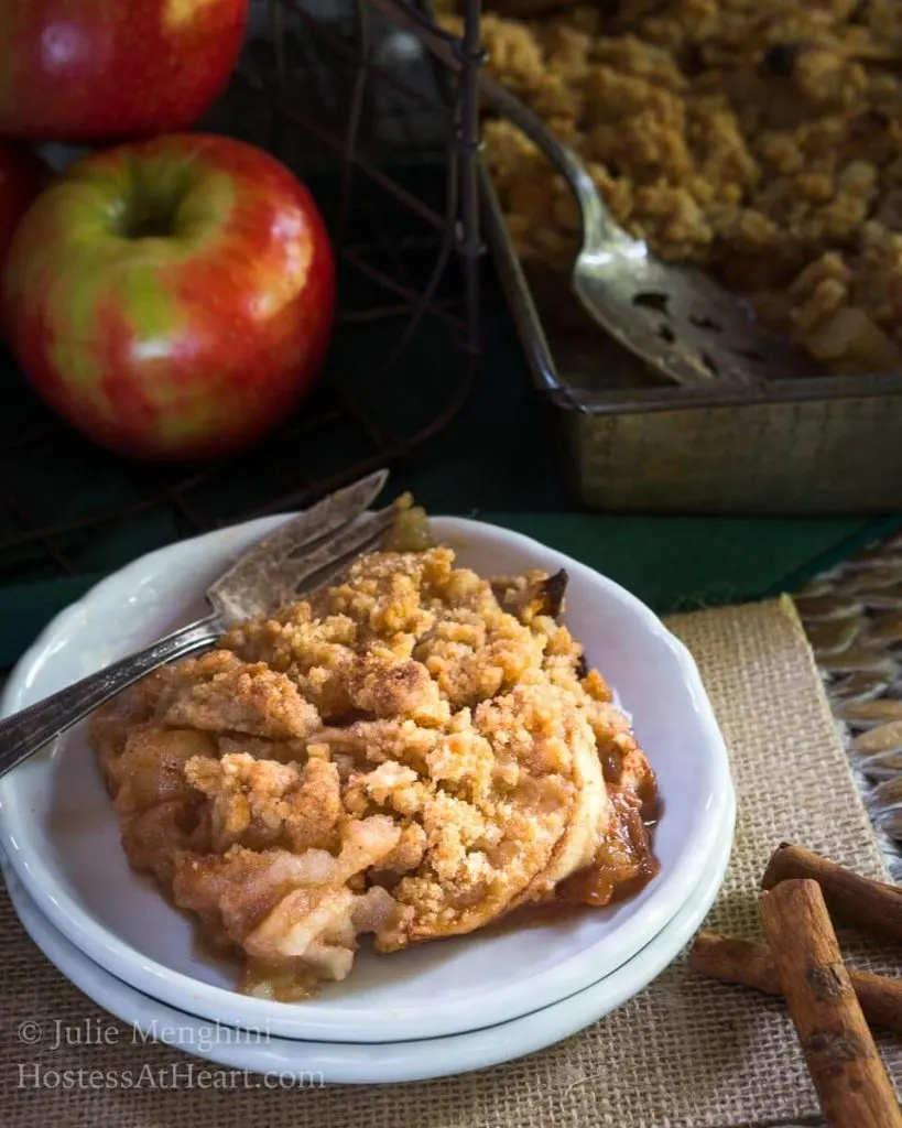Top angled view of a slice of apple crisp sitting on a stack of two white plates over a piece of burlap. The pan of crisp sits in the background next to fresh apples and cinnamon sticks.