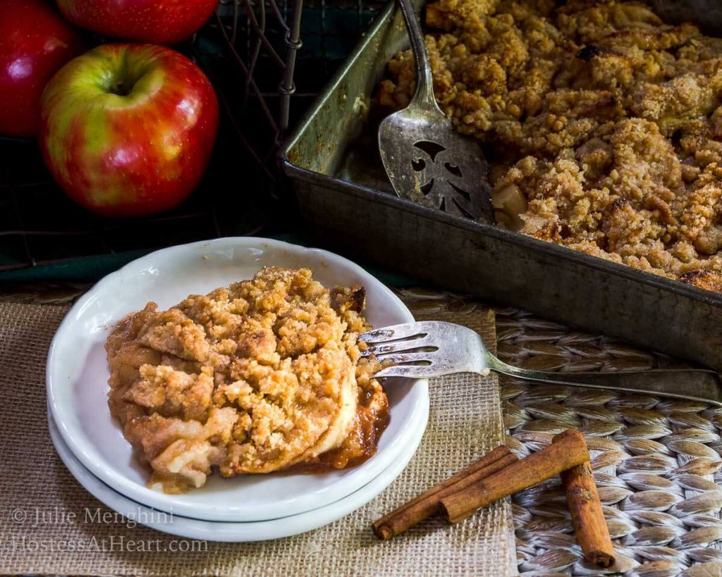 Top angled view of a slice of apple crisp sitting on a stack of two white plates over a piece of burlap. The pan of crisp sits in the background next to fresh apples and cinnamon sticks.