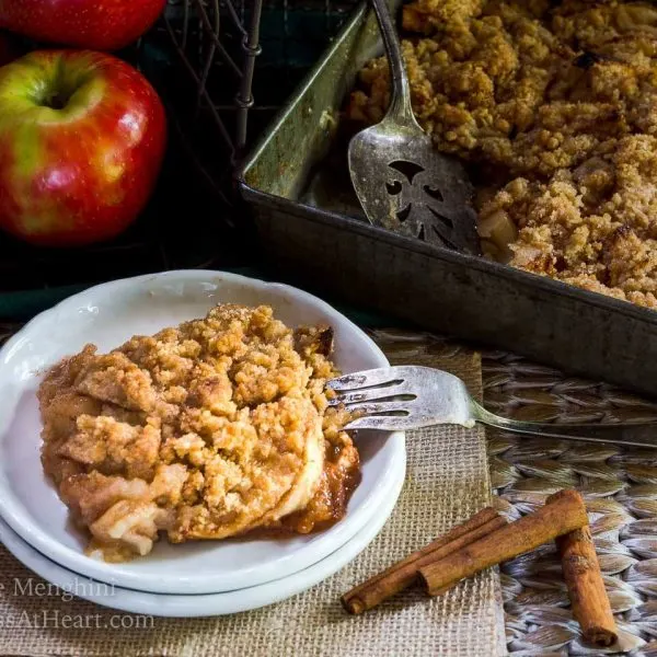 Top angled view of a slice of apple crisp sitting on a stack of two white plates over a piece of burlap. The pan of crisp sits in the background next to fresh apples and cinnamon sticks.