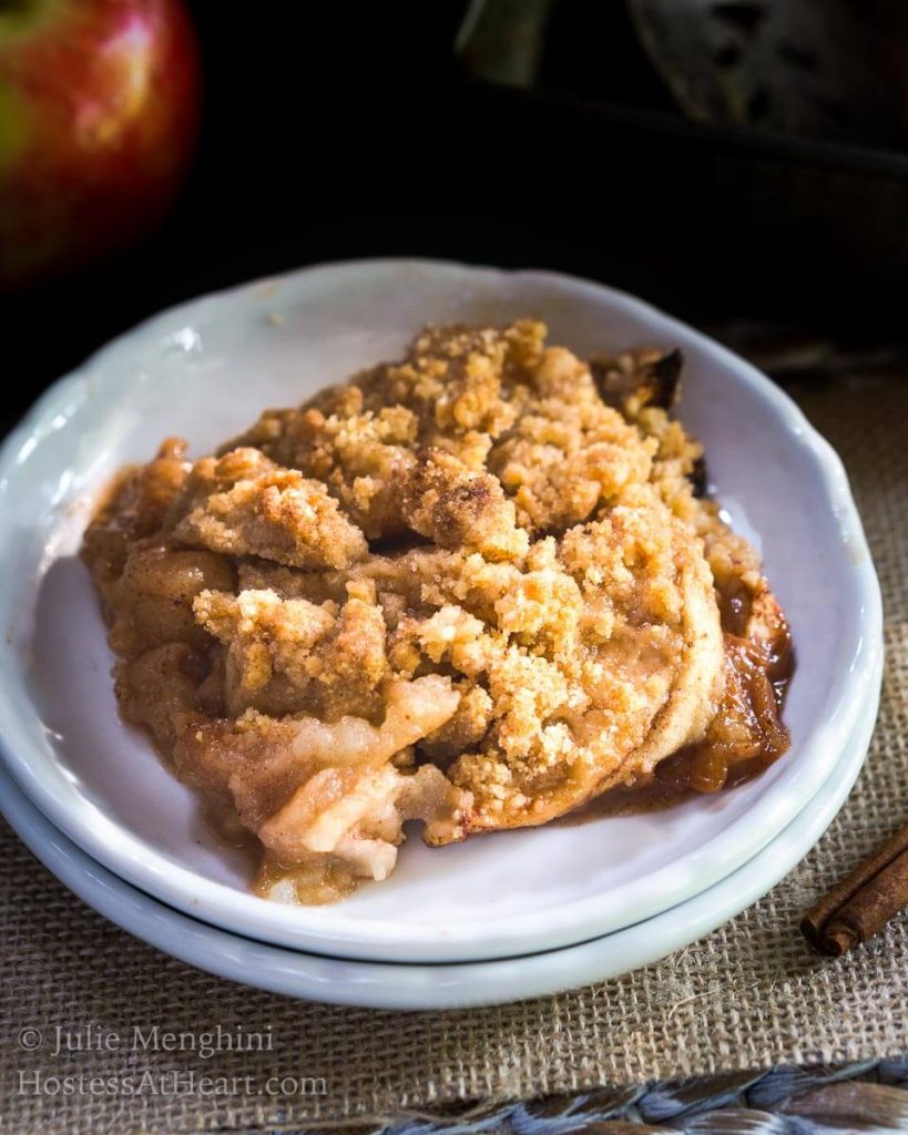 Top angled view of a slice of apple crisp sitting on a stack of two white plates over a piece of burlap. Cinnamon sticks sit next to the plates.