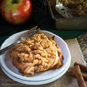 A top-down view of apple crisp with a crumble top sitting on a stack of two plates. A fork sits next to the apple crisp and a fresh apple sits in the background next to the pan of apple crisp.
