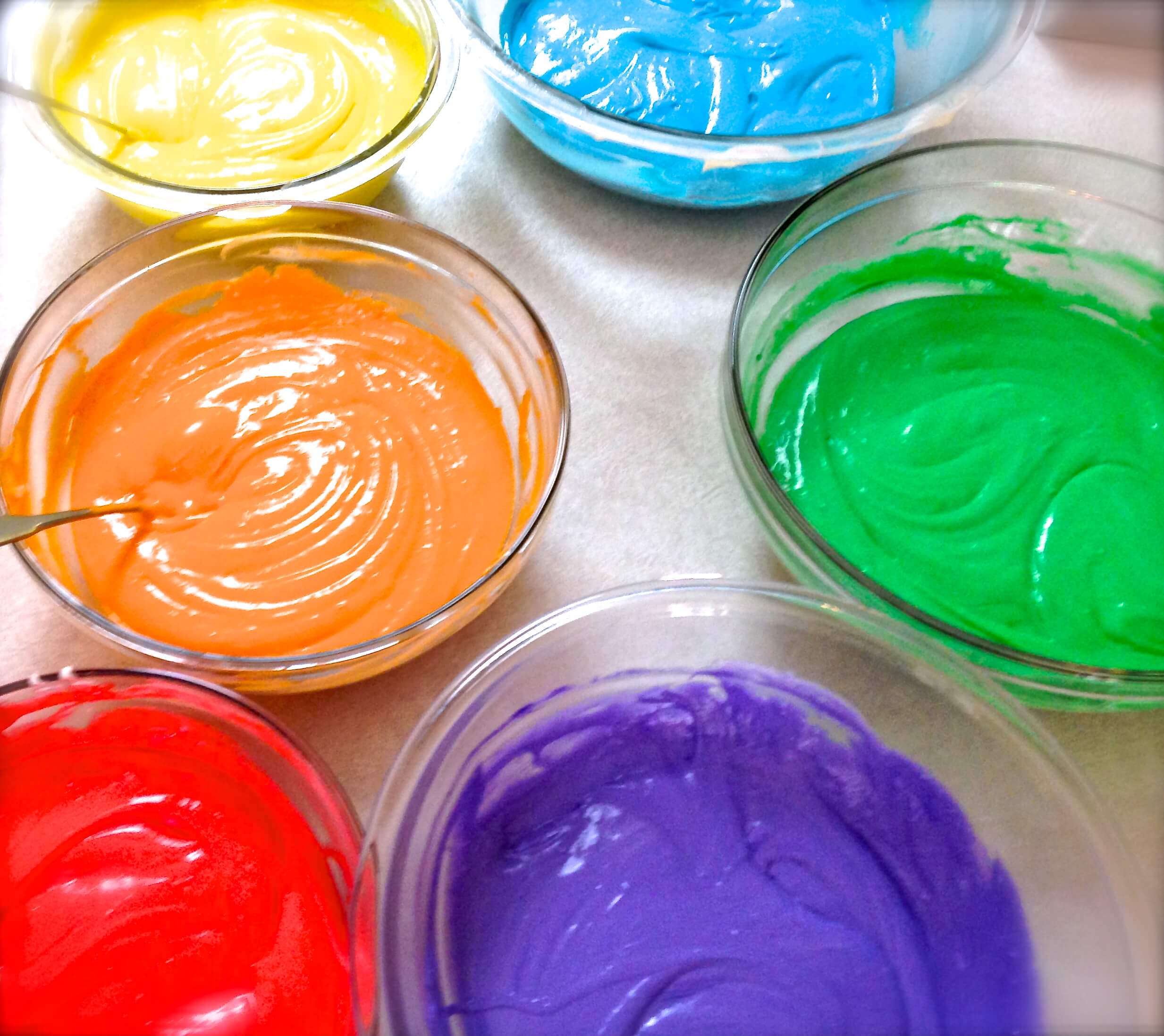 Bowls of dyed cake batter in blue, orange, yellow, green, purple, and red.