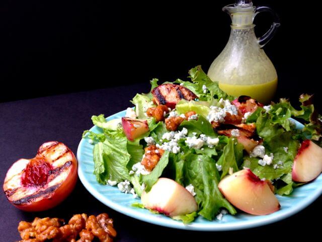 A light blue plate holding a green lettuce salad tossed with grilled peaches. A carafe of Honey Jalapeno dressing, a grilled peach half,  and toasted walnuts sit next to the plate.