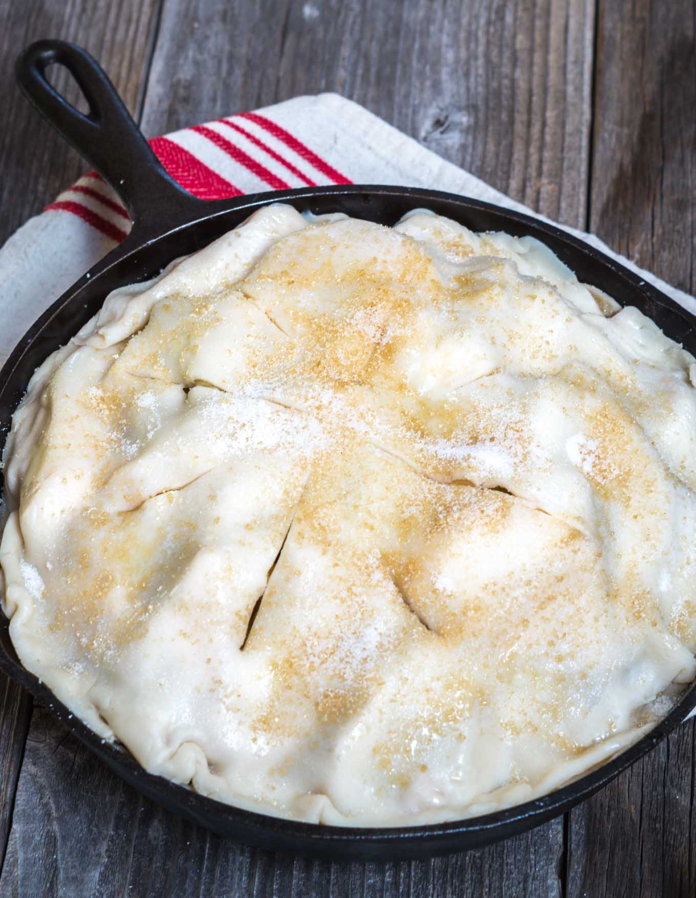 Unbaked Apple Pie in a cast-iron skillet dusted with sugar and sliced to vent the apple pie sitting on a red striped napkin.