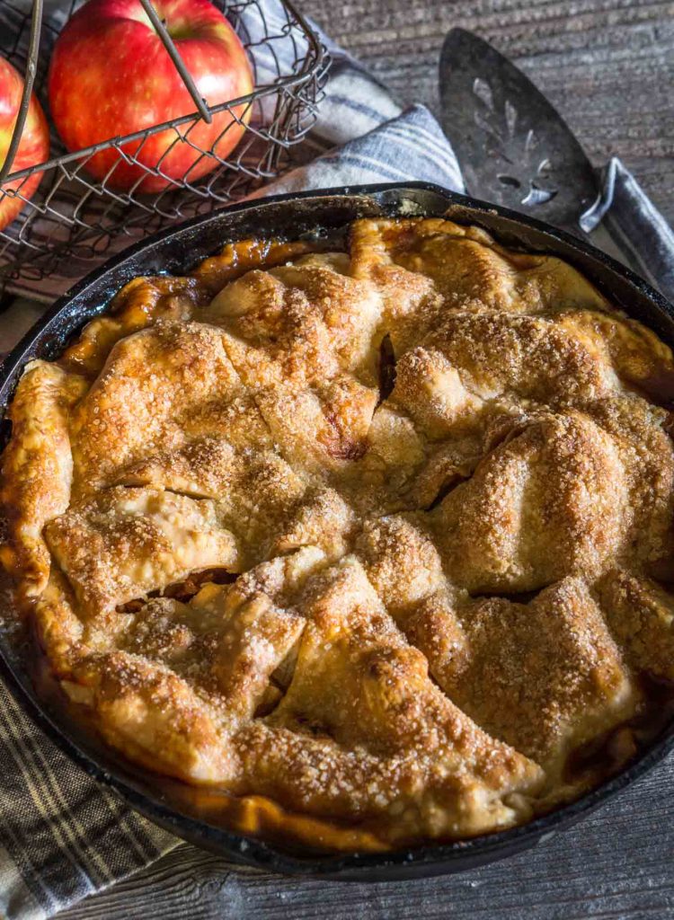 Draufsicht auf 3/4 eines Apfelkuchens, der in einer gusseisernen Pfanne gebacken wird, mit einem Korb frischer Äpfel dahinter