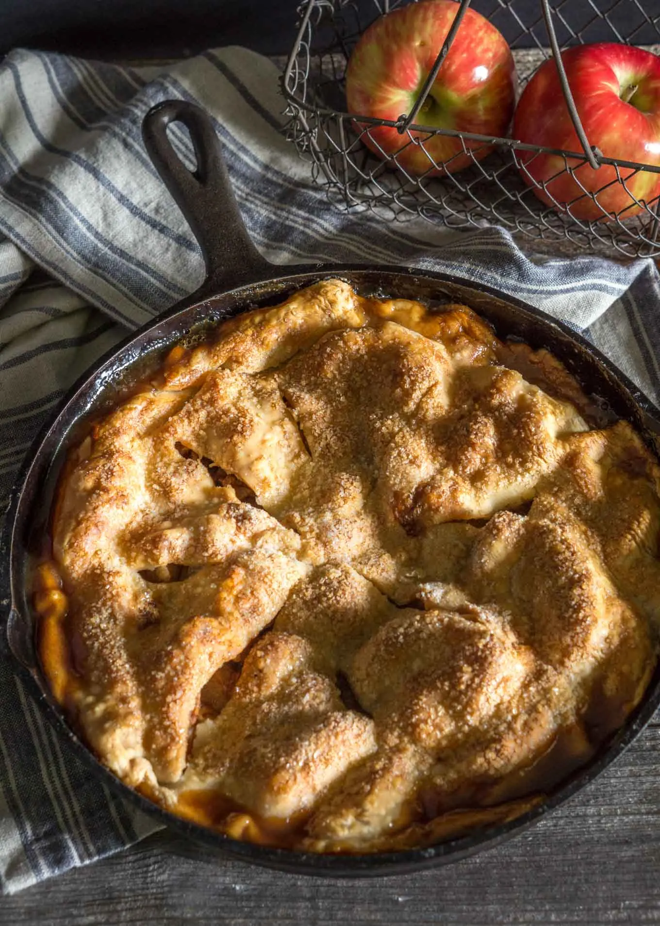Top view of a Skillet Apple Pie in a cast iron skillet sitting in front of a basket of apples on a blue towel covering a rustic wood background.