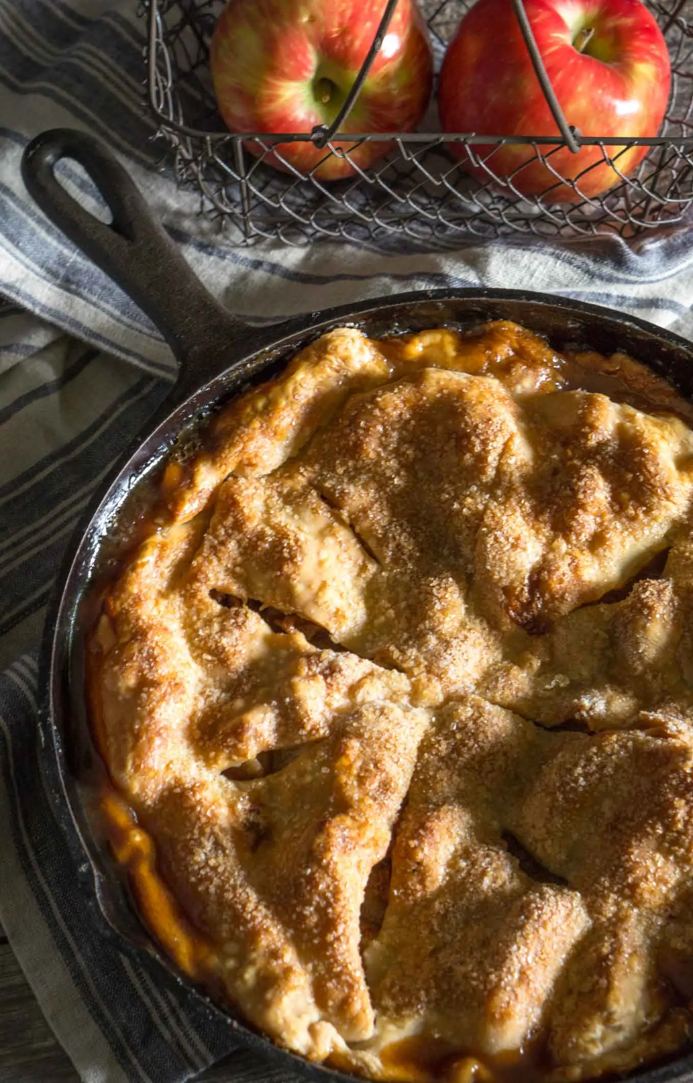 Close up of a baked apple pie sitting in a cast iron skillet on top of a blue stripped towel in front of a basket of red apples.