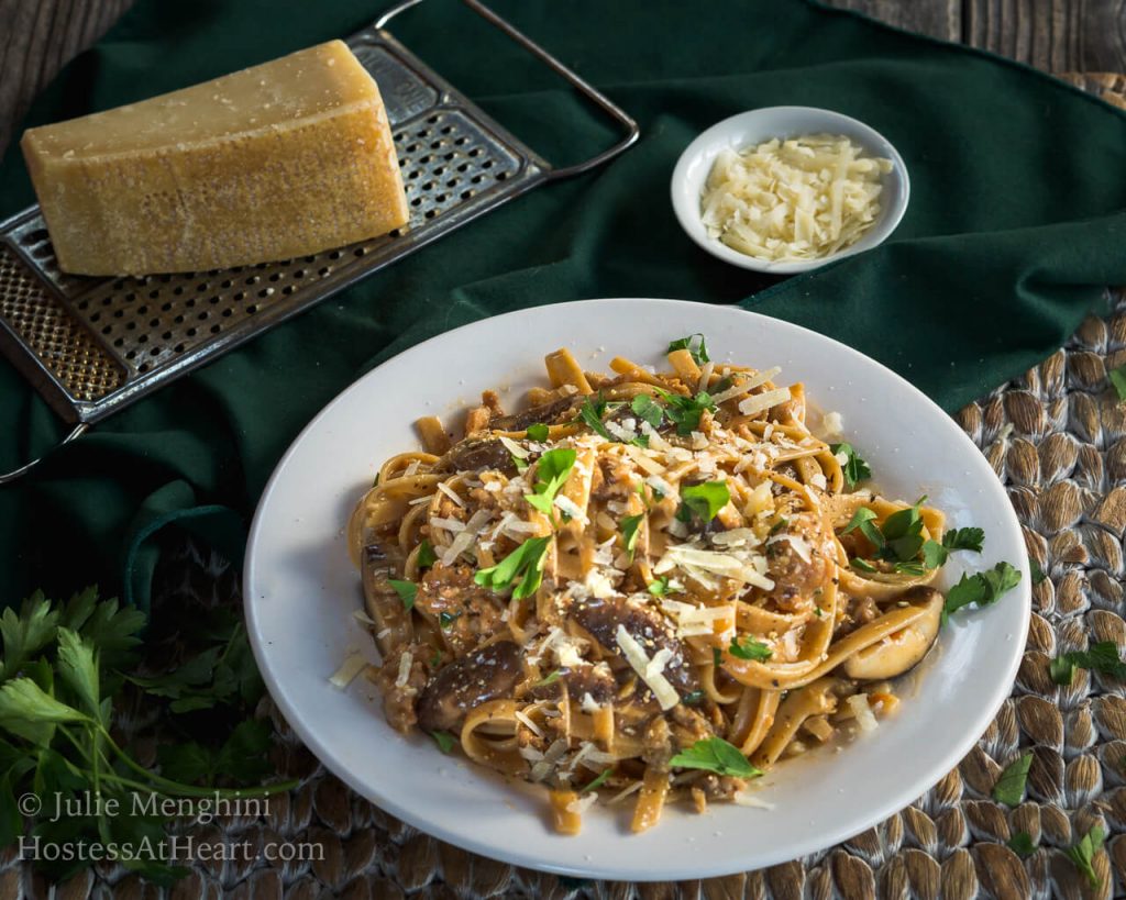 Top view of a white plate filled with Fettuccine pasta combined with sausage, mushrooms, and cheese and garnished with fresh parsley. A block of parmesan sits over a grater next to a wire dish of grated cheese over a green napkin.