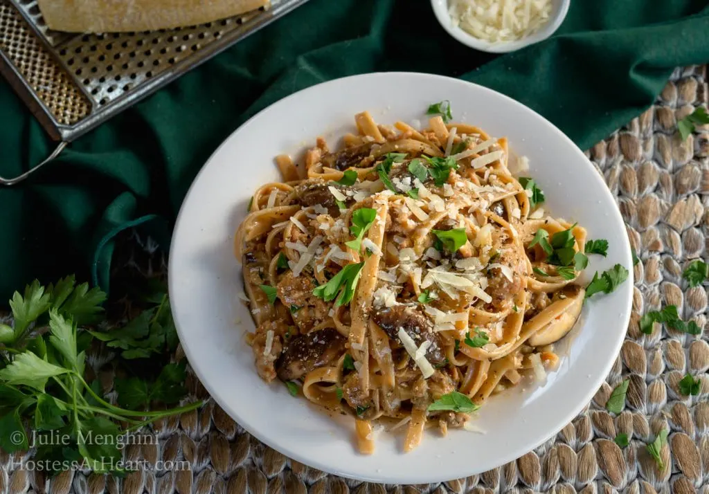 Top-down view of a white plate filled with Fettuccine pasta combined with sausage, mushrooms, and cheese and garnished with fresh parsley. A block of parmesan sits over a grater next to a wire dish of grated cheese over a green napkin.