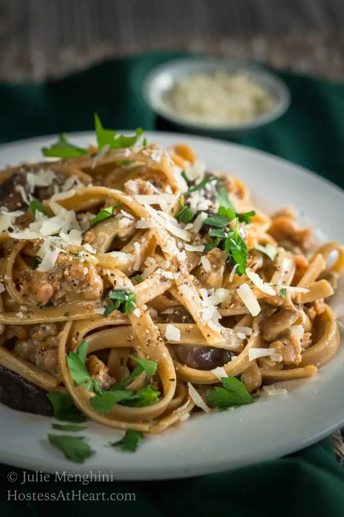 Close up side view of a white plate filled with Fettuccine pasta combined with sausage, mushrooms, and cheese and garnished with fresh parsley. A block of parmesan sits over a grater next to a wire dish of grated cheese over a green napkin.