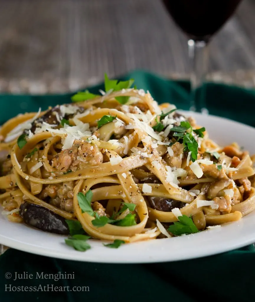 Side view of a white plate filled with Fettuccine pasta combined with sausage, mushrooms, and cheese and garnished with fresh parsley. A block of parmesan sits over a grater next to a wire dish of grated cheese over a green napkin. A glass of wine sits in the background.