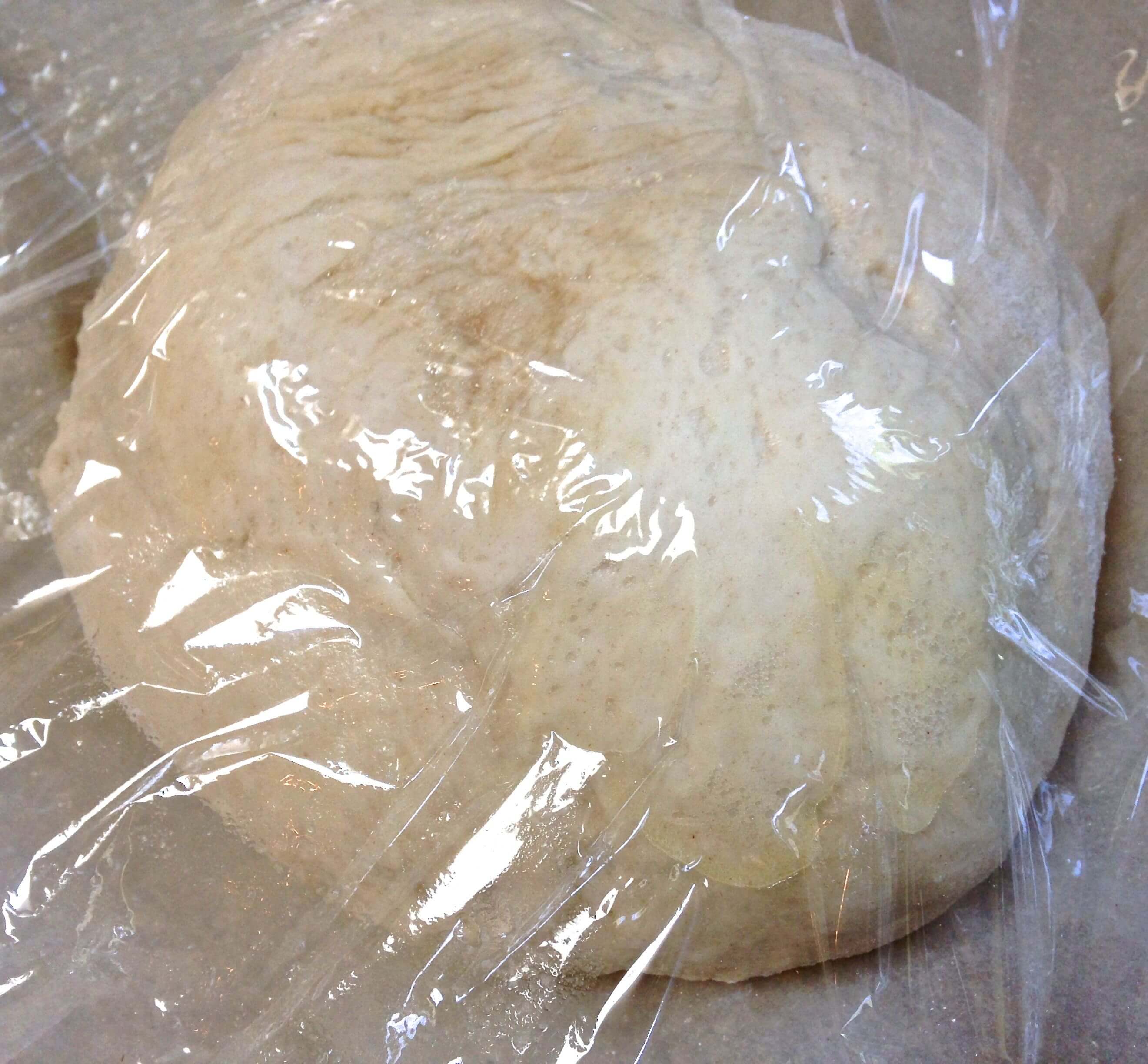 A ball of raw bread dough sitting in a glass bowl covered with plastic wrap.