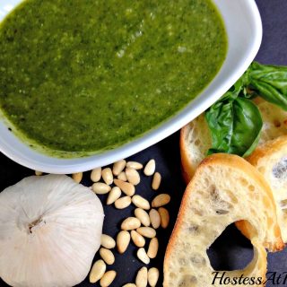 Top-down view of pesto sitting in a white bowl. Slices of a baguette, garlic, pinenuts, and sweet basil sit to the side.