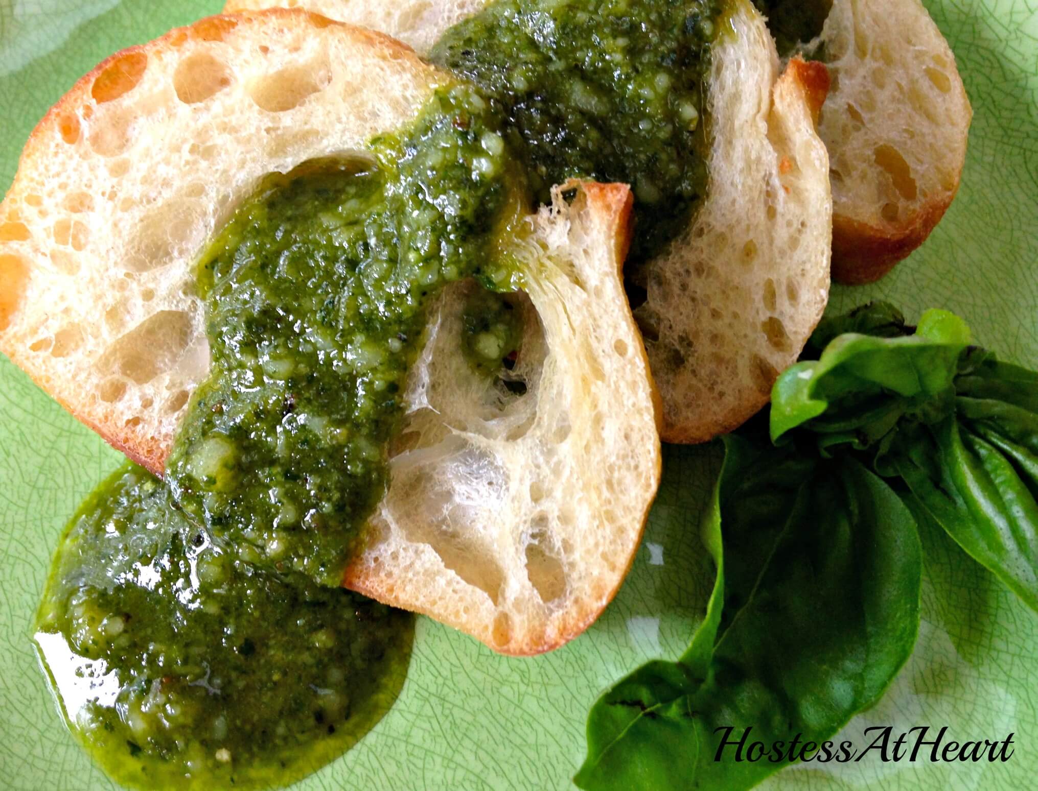 Two views of Top-down view of pesto sitting in a white bowl. Slices of a baguette, garlic, pinenuts, and sweet basil sit to the side.