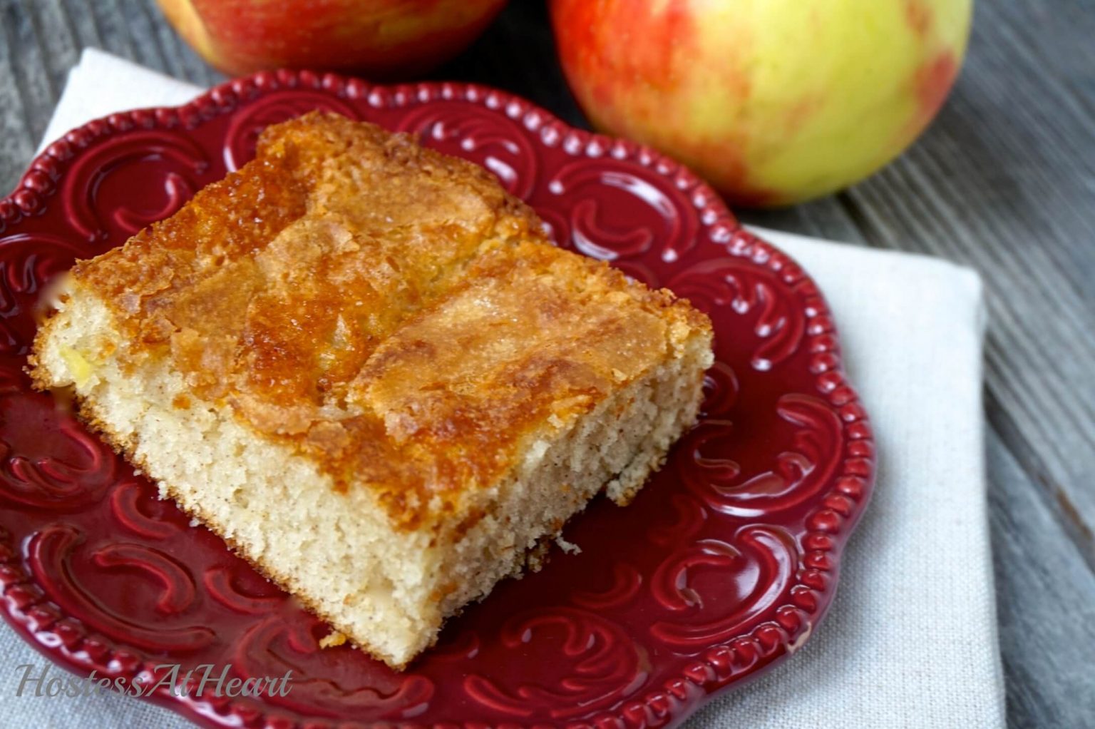 A piece of Apple cake sitting on a red plate. Fresh apples are in the background.