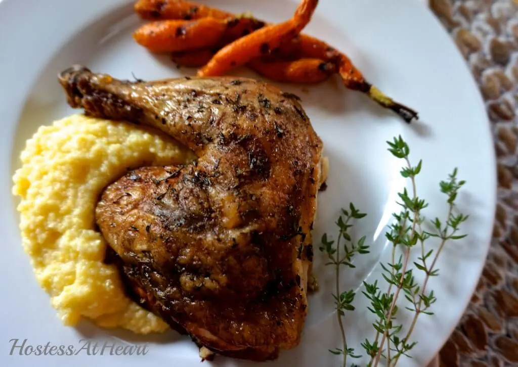 Top-down view of a grilled chicken quarter laying on a bed of polenta on a white plate with a garnish of fresh thyme.