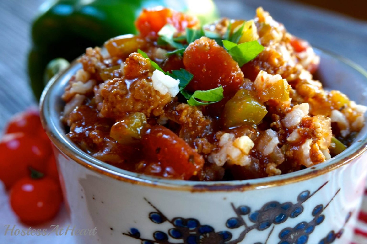 Sideview of a soup bowl with a blue design on the front is heaped with Stuffed Pepper Sour that\'s full of sausage, peppers, tomatoes, and garnished with fresh parsley. Fresh peppers and tomatoes sit in the background.