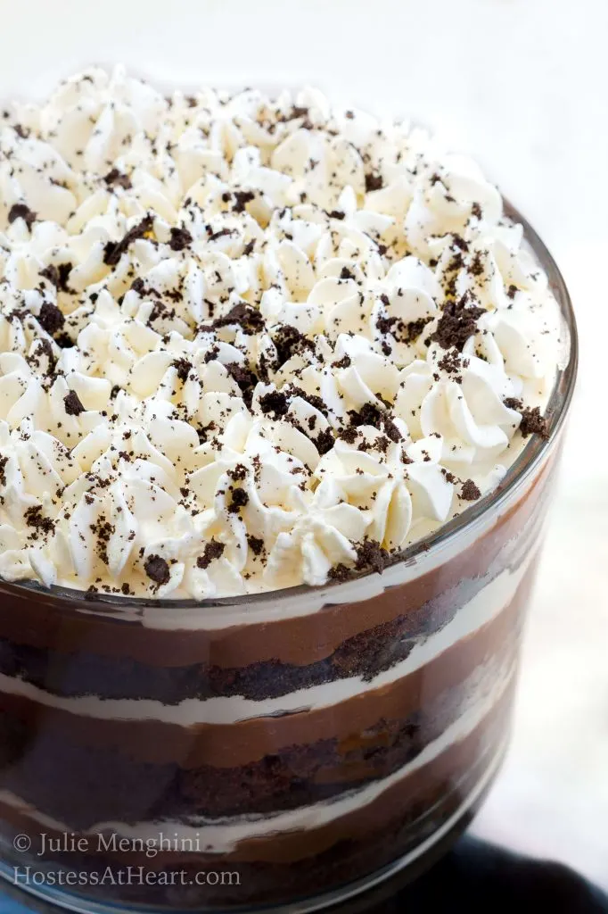 Top-angle of piped whipped cream with crumbles of oreo cookies sprinkled over the top. The layers of chocolate pudding, whipped cream, and chocolate cake show through the side of the glass trifle bowl.