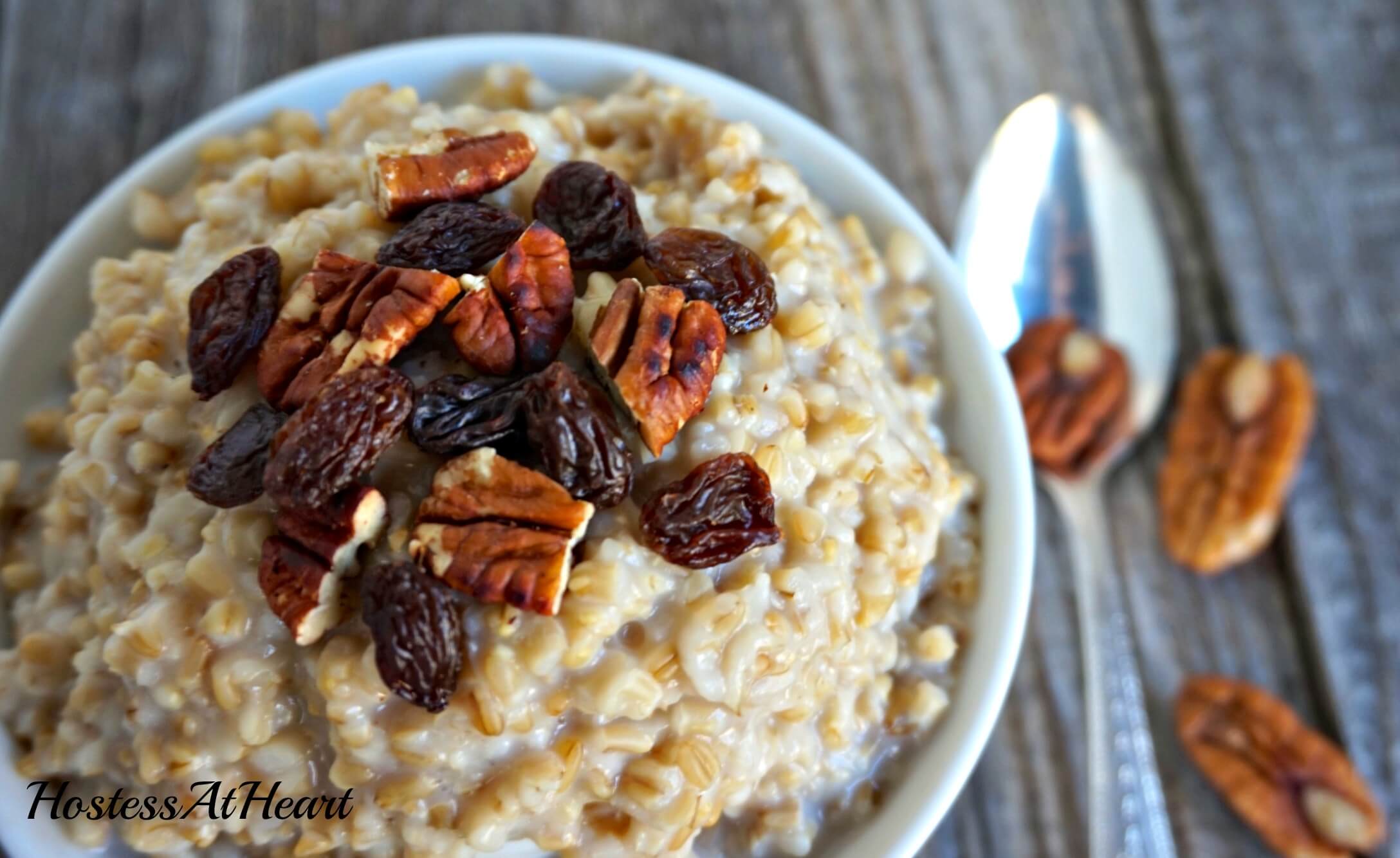 A white bowl filled with Steel Cut Oatmeal and garnished with raisins and chopped pecans. A spoon with a nut on it sits to the side.