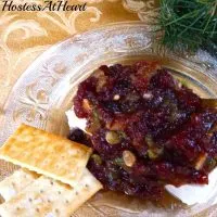 A glass plate topped with a brick of cream cheese that's topped with a Cranberry Pistachio sauce. Crackers sit off to the side.