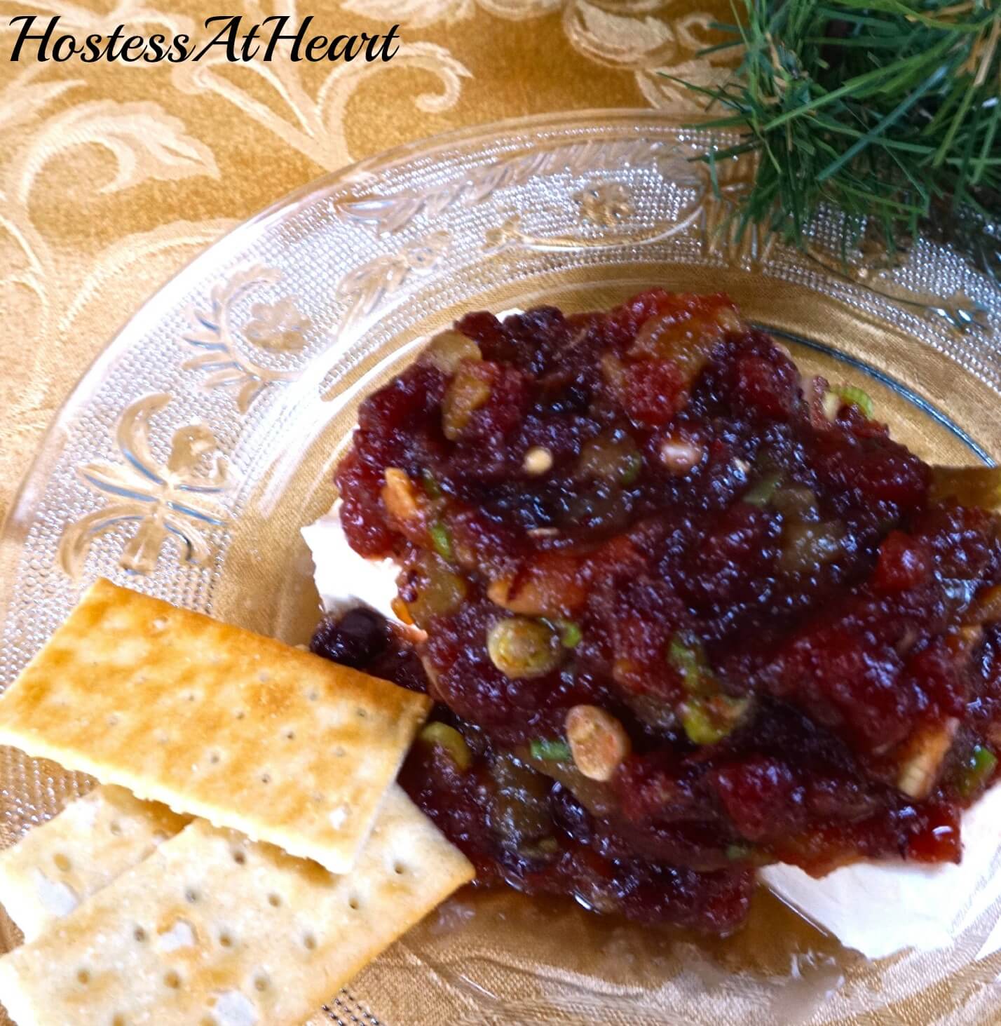 A glass plate topped with a brick of cream cheese that\'s topped with a Cranberry Pistachio sauce. Crackers sit off to the side.