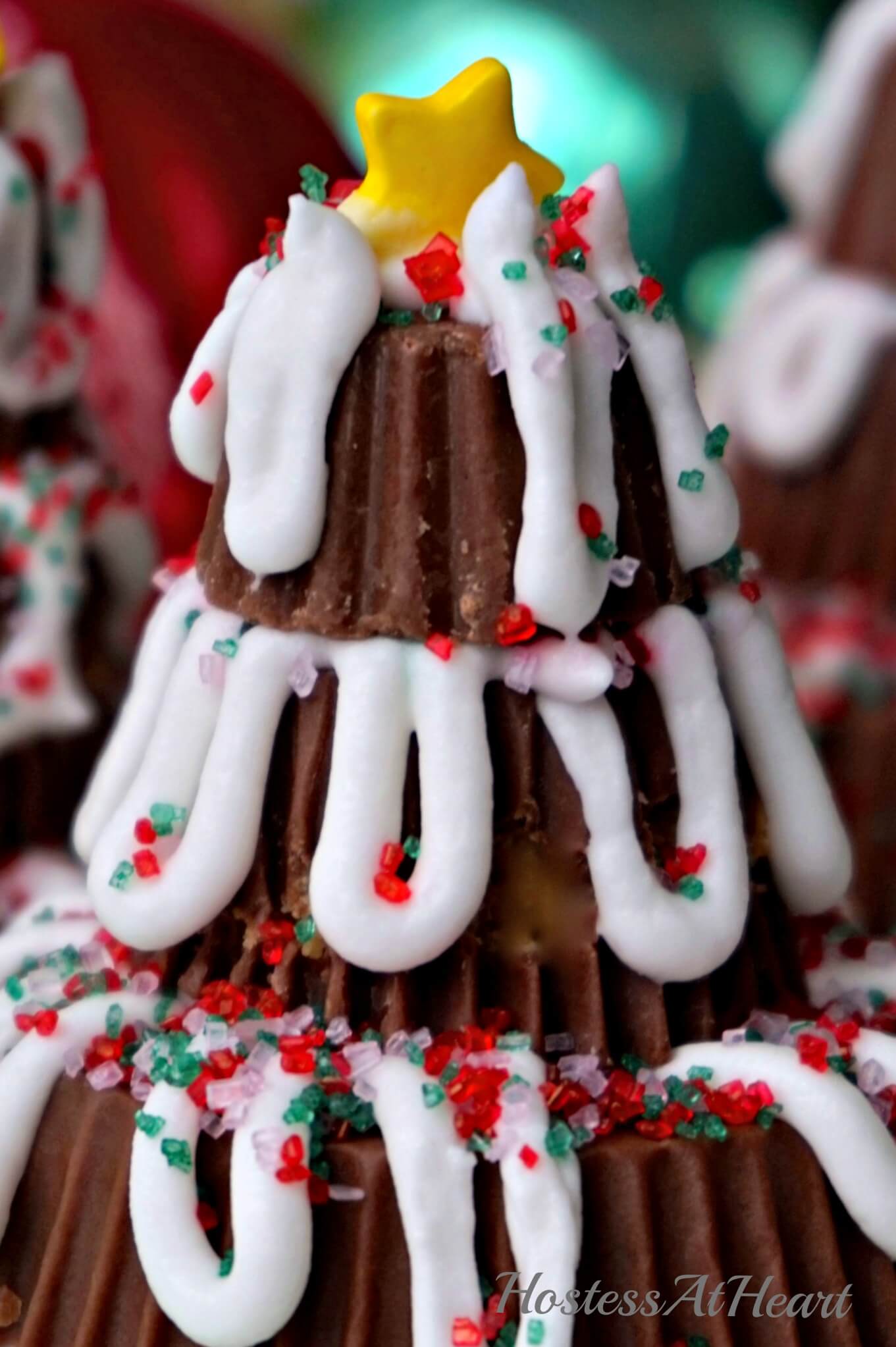 A Christmas tree made from Reeses Peanut Butter cups decorated with frosting and sprinkles with Ornaments in the background.