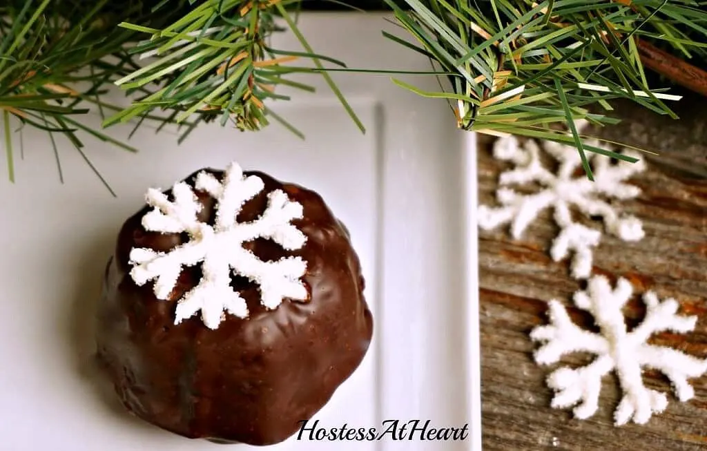 A white plate holding a small cake bundtlette frosted with chocolate ganache and topped with a sugar snowflake. 