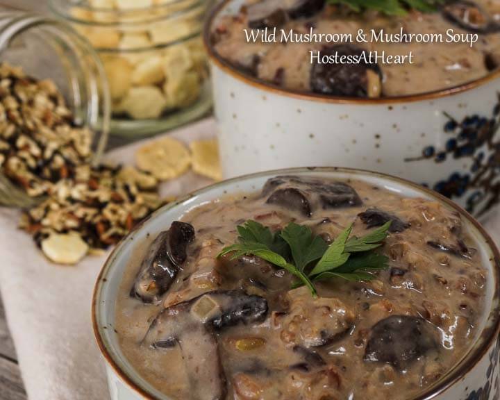 Two soup bowls filled with Wild Rice and Mushroom soup topped with fresh parsley. Soup crackers are scattered about and an antique soup spoon sits to the side. Jars of soup crackers and while rice sit in the background.