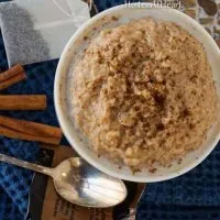 Top-down photo of a white bowl filled with Chai-Spiced Steel Cut Oatmeal. Cinnamon sticks, a spoon, and a chai tea bag sit next to the bowl.