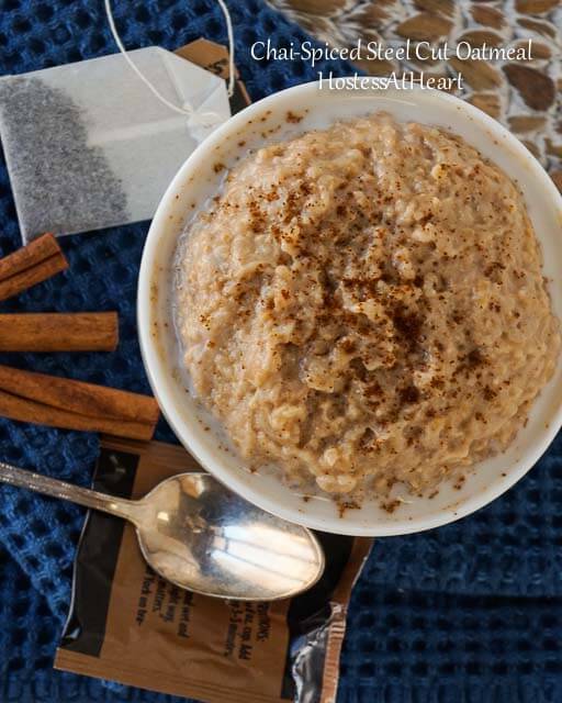 Top-down photo of a white bowl filled with Chai-Spiced Steel Cut Oatmeal. Cinnamon sticks, a spoon, and a chai tea bag sit next to the bowl.