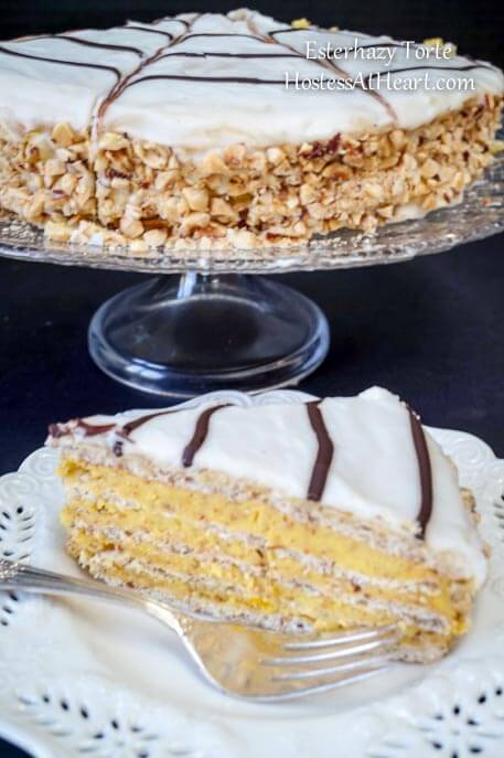 An Esterhazy torte with a spiderweb design over the top and the sides pressed with crushed nuts on a cake plate with a slice of the cake in the front showing the layers of the nutty cream filling.