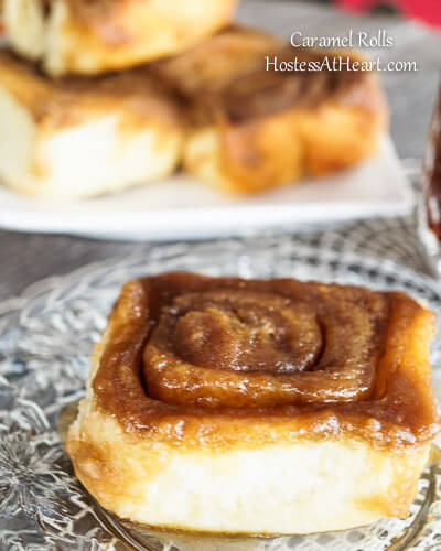 A caramel roll sitting on a glass plate with more rolls stacked on a plate in the background next to a cup of coffee.