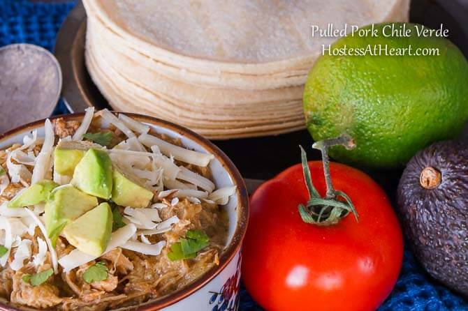 A bowl Pulled Pork Green Chile Verde surrounded by fresh lime, tomato, avocado, and tortillas.