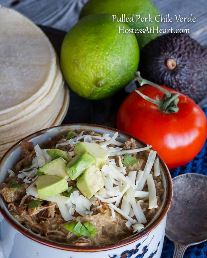 A bowl Pulled Pork Green Chile Verde surrounded by fresh lime, tomato, avocado, and tortillas.