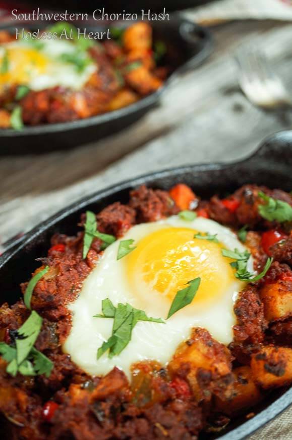 A cast-iron dish filled with Chorizo Hash and topped with a poached egg. A fork full of the hash hovers over the dish and the second dish of hash sits in the background.