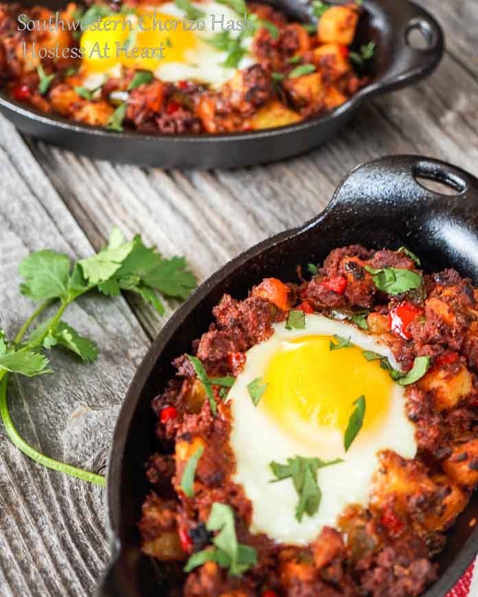 A cast-iron dish filled with Chorizo Hash and topped with a poached egg. A fork full of the hash hovers over the dish and the second dish of hash sits in the background.