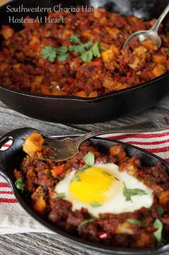 A cast-iron dish filled with Chorizo Hash and topped with a poached egg. A fork full of the hash hovers over the dish and A cast-iron pan of hash sits in the background.