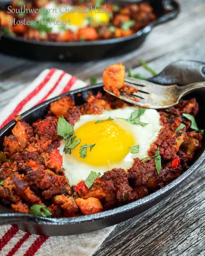 A cast-iron dish of Chorizo Hash topped with a poached egg. A second dish sits in the background over a wooden background.