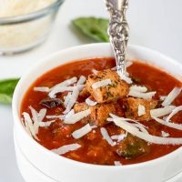 A white bowl of tomato Florentine soup garnished with shredded cheese and croutons with a spoon sitting in the soup A small bowl of cheese sits in the background. 