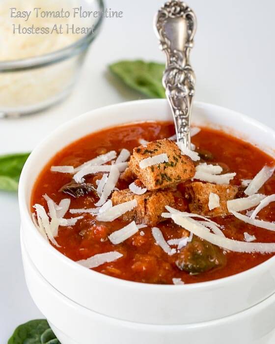 A white bowl of tomato Florentine soup garnished with shredded cheese and croutons with a spoon sitting in the soup A small bowl of cheese sits in the background.  